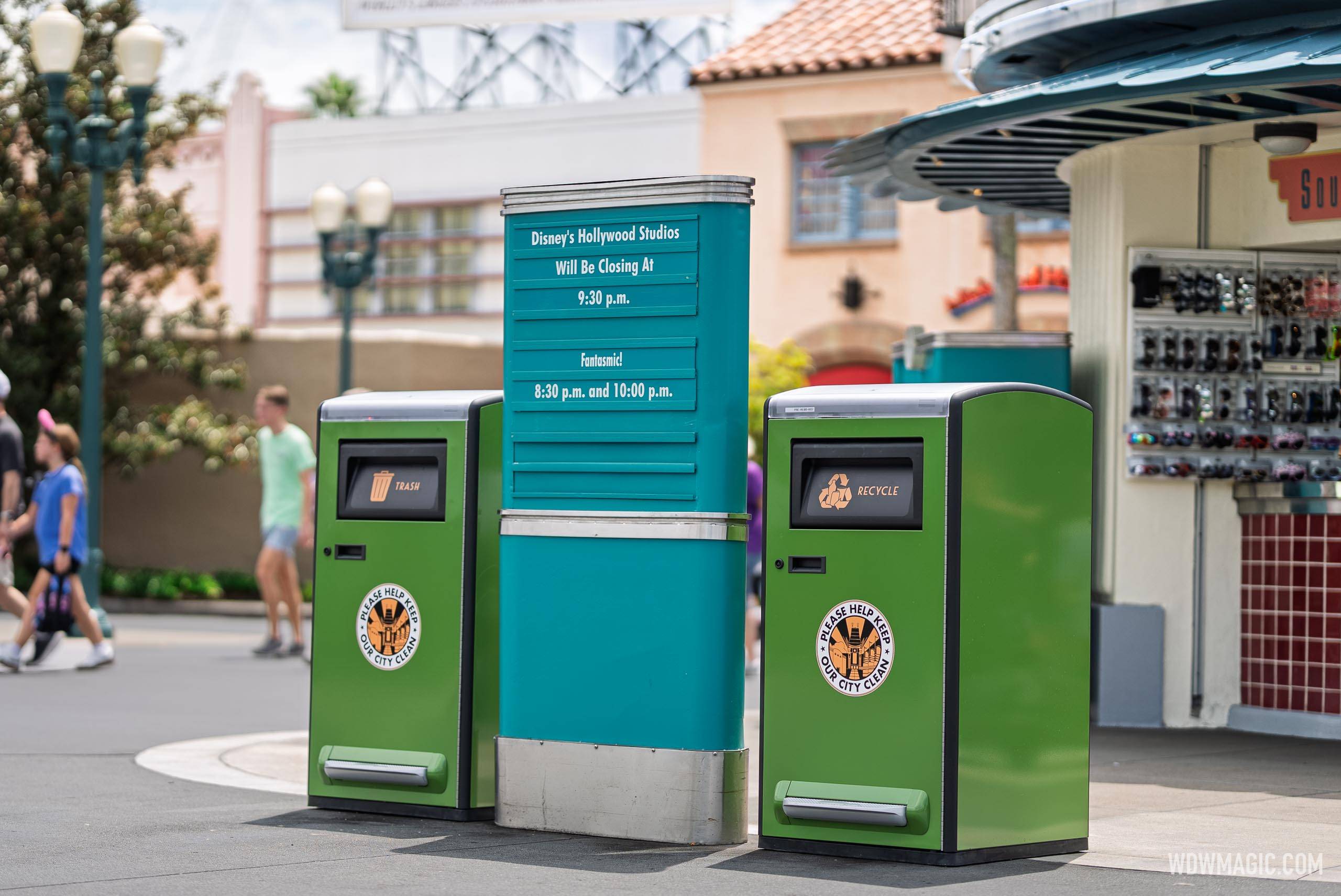 New green solar-powered trash and recycling bins flank a park information board showing closing times for Fantasmic! at Disney's Hollywood Studios
