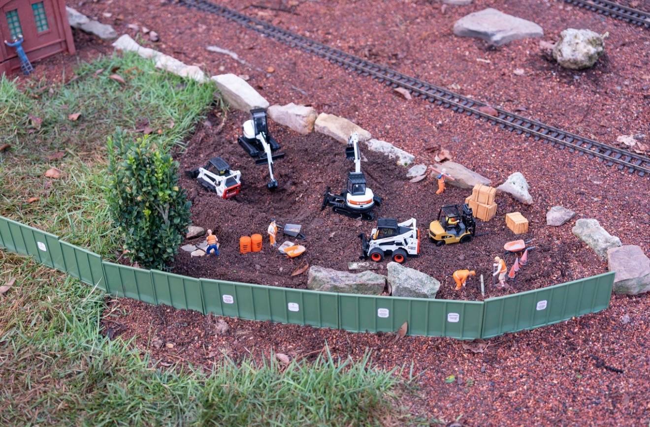 Miniature Bobcat skid steers, excavators, a forklift, and construction worker figures populate a small dirt work site within the Germany pavilion train garden