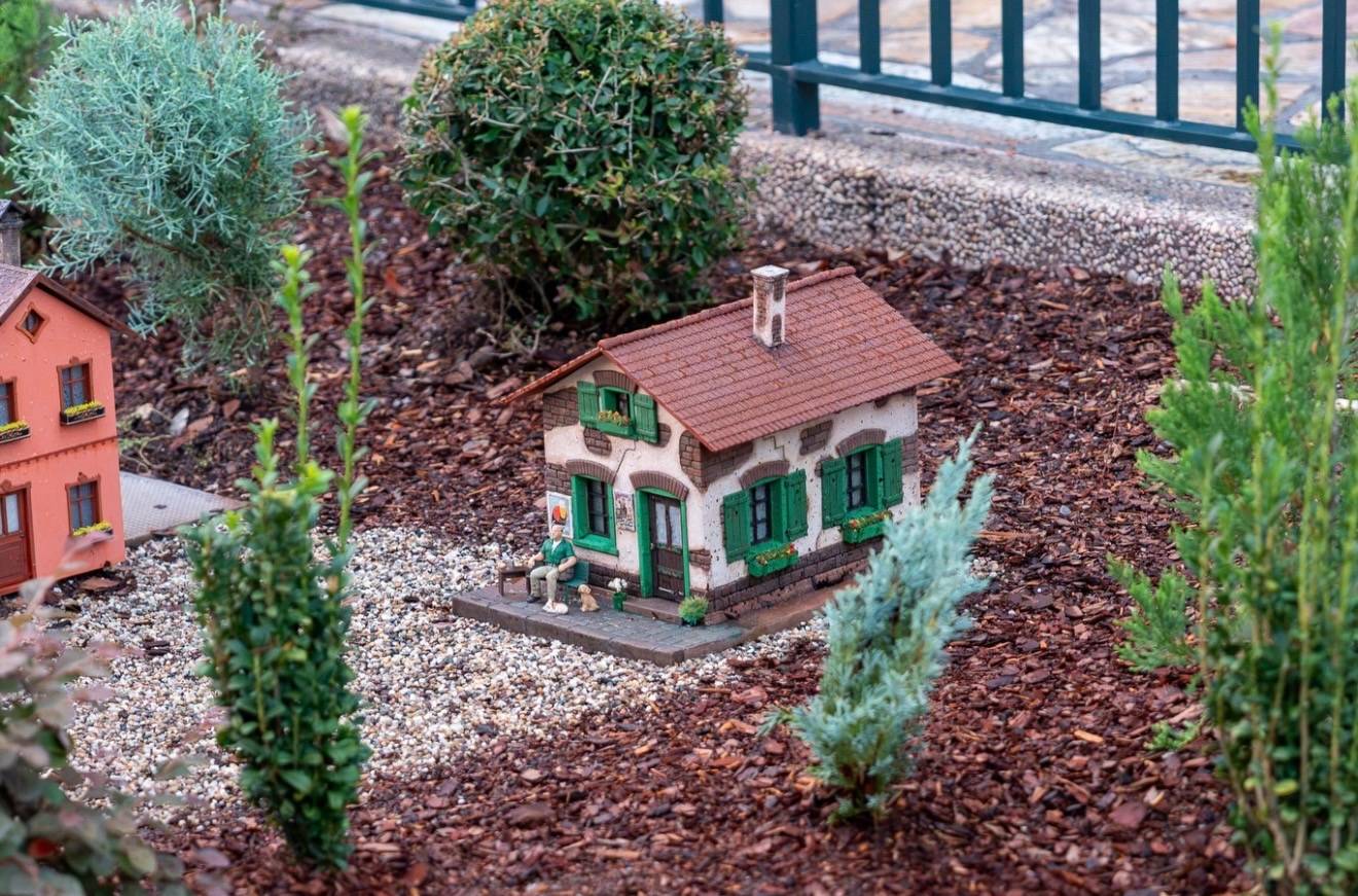 Miniature white cottage with green shutters and a chimney sits among small-scale figures, gravel, and topiaries in the Germany pavilion train garden