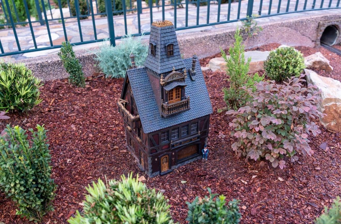 Dark Gothic-style miniature building with a pointed tower, decorative balcony, and a small figurine standing at its entrance in the Germany pavilion train garden