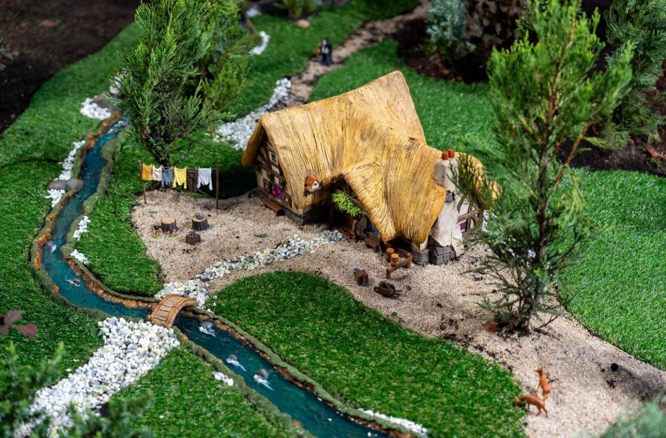 Thatched-roof cottage with a clothesline, miniature deer, and a winding stream crossed by a small wooden bridge in the Germany pavilion train garden