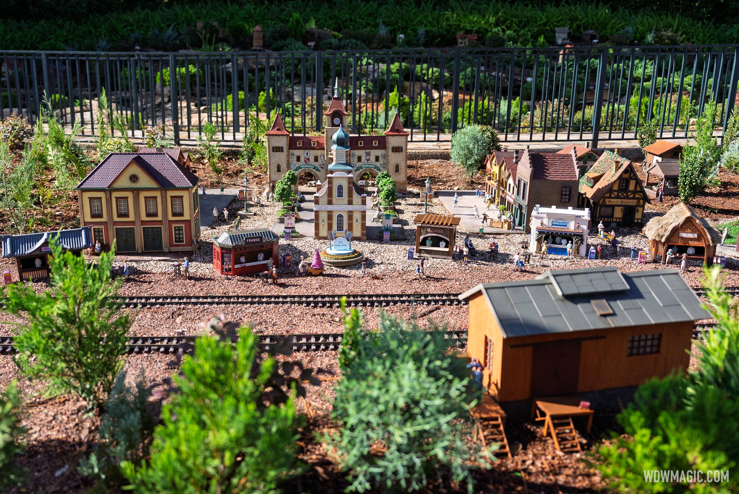The Germany pavilion train garden overview, showing the France and Greece Food & Wine Festival booths, a central fountain, and European-style buildings with miniature figures throughout
