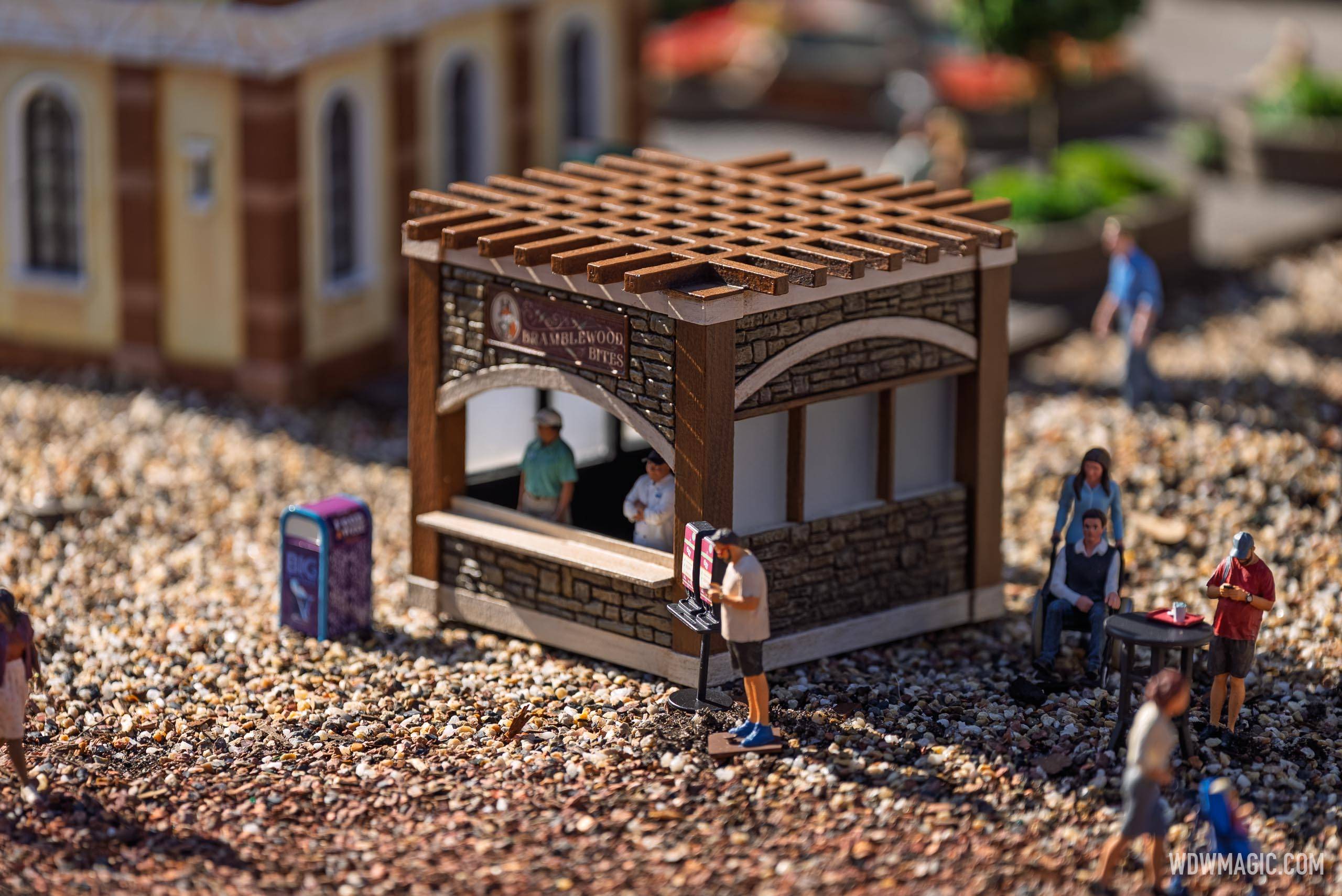 The Bramblewood Bites booth miniature in the Germany pavilion train garden features tiny figures at the counter and a menu board outside, set among pebble-covered ground