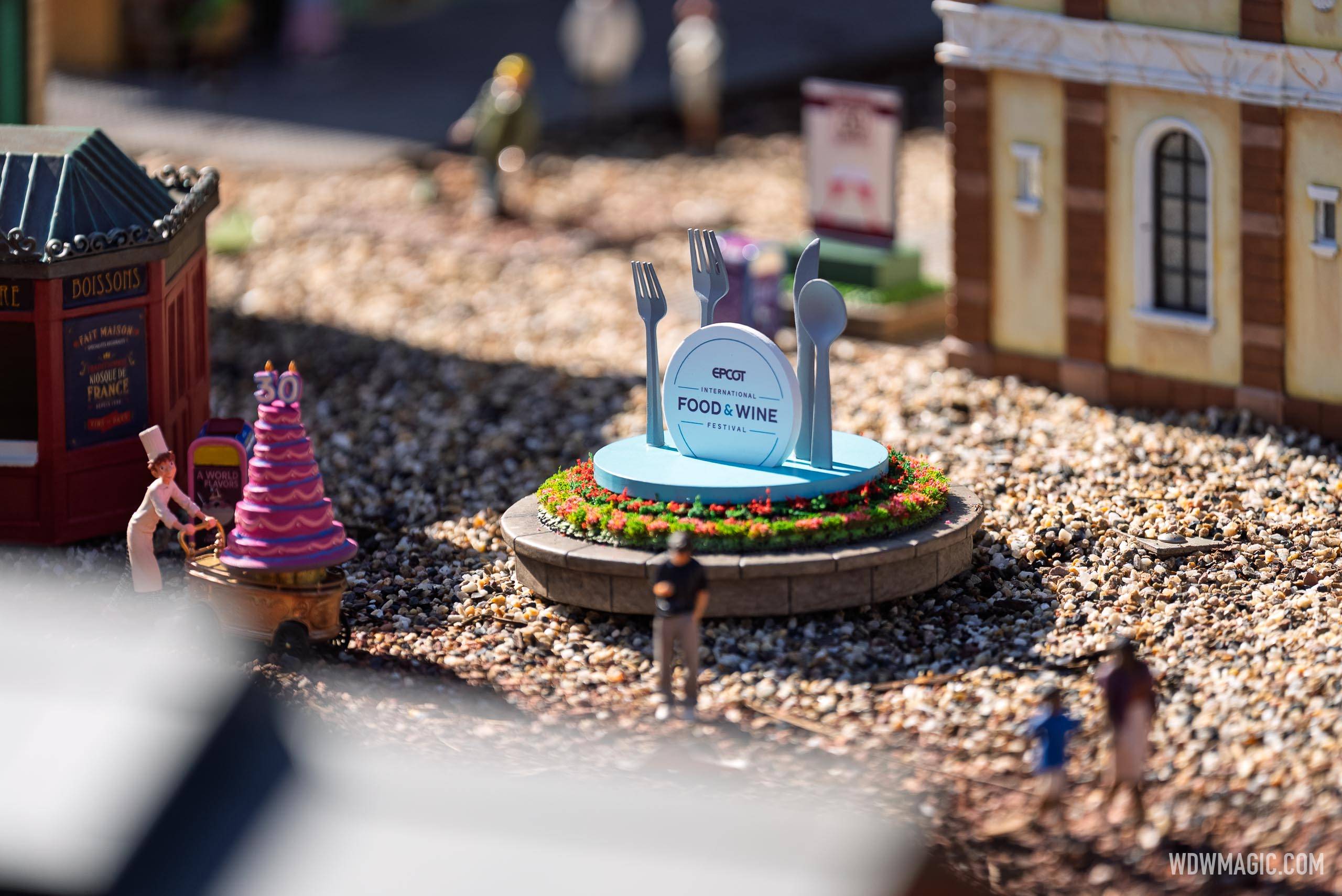 The EPCOT International Food & Wine Festival sign sits on a circular planter surrounded by miniature figures, a France booth, and a tiered celebration cake display in the Germany train garden