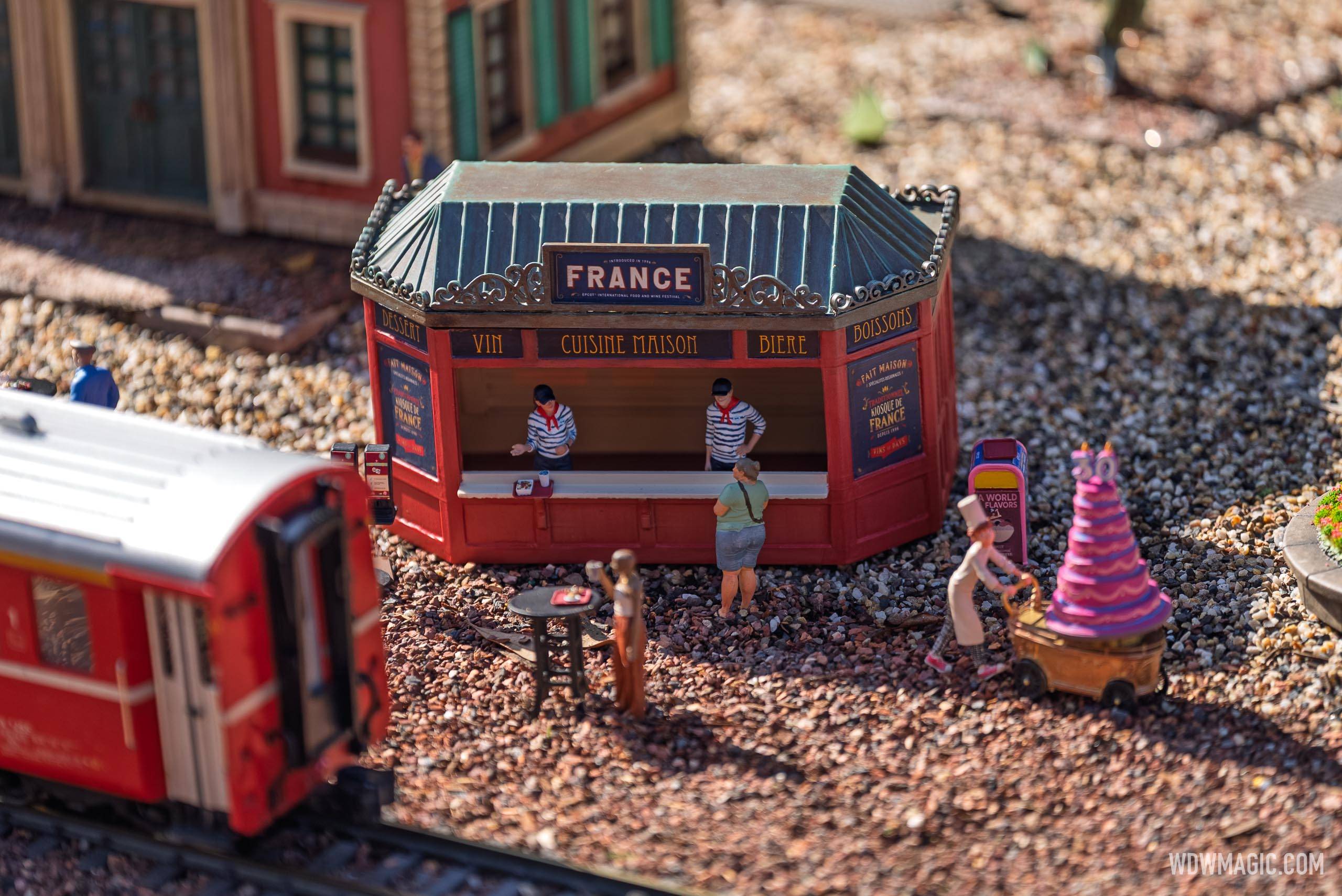 The France booth miniature in the Germany pavilion train garden features tiny figures at the counter, with signage for Vin, Cuisine Maison, Bière, and Boissons