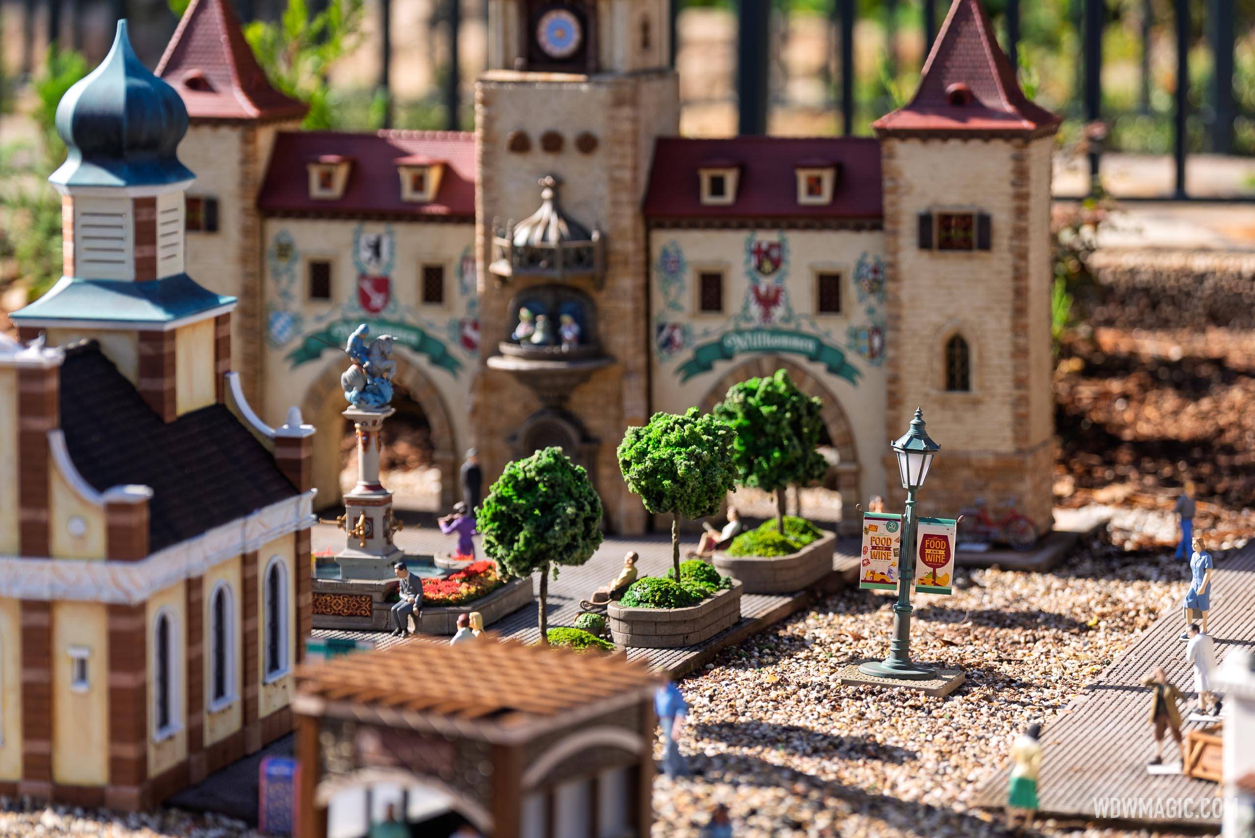 The Germany pavilion train garden features a detailed miniature of the Willkommen gate, clock tower, and central plaza with tiny trees, figures, and Food & Wine Festival signage on a lamp post