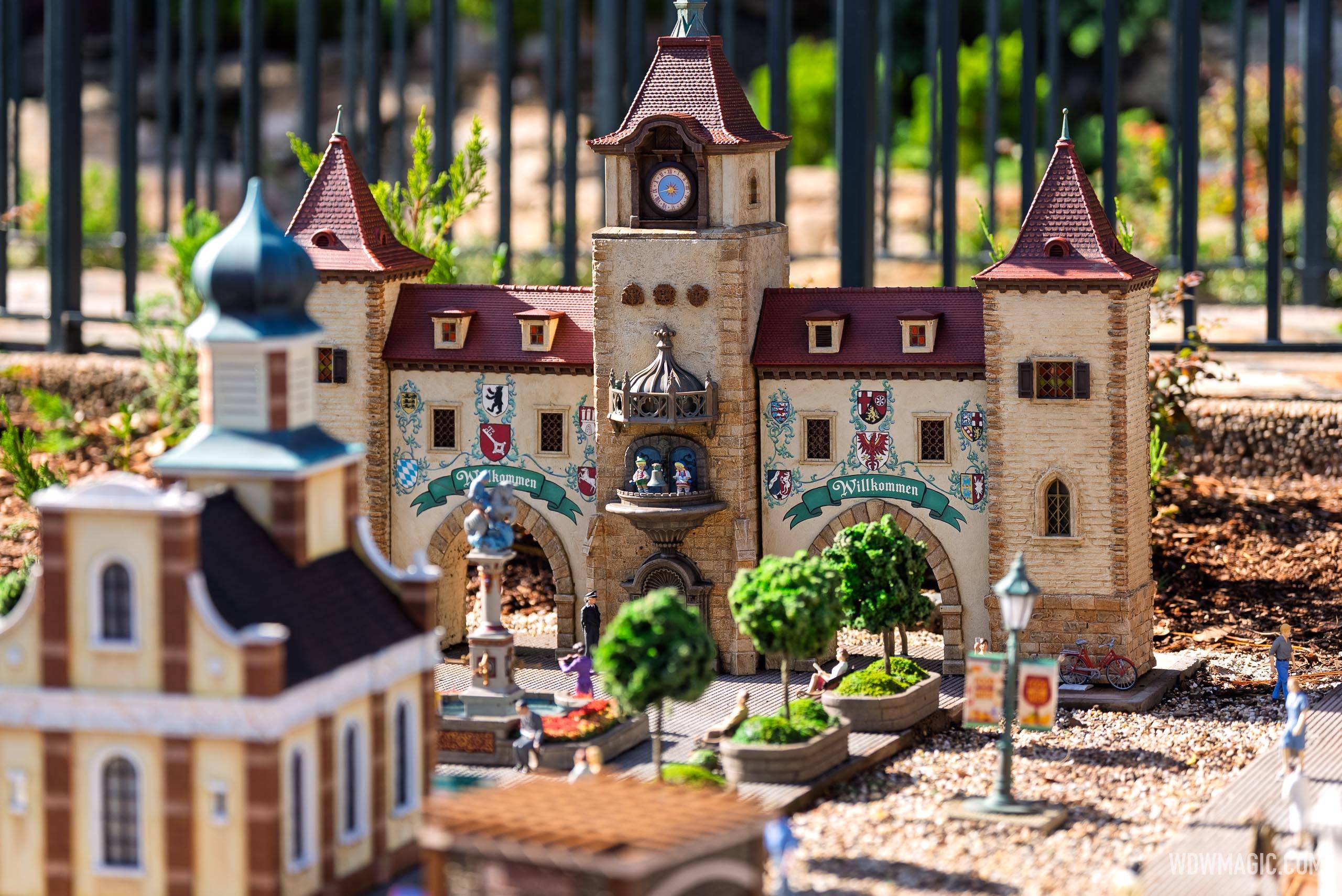 The Willkommen gate and clock tower miniature in the Germany pavilion train garden, surrounded by tiny topiary trees, lamp posts, and miniature figures in the central plaza