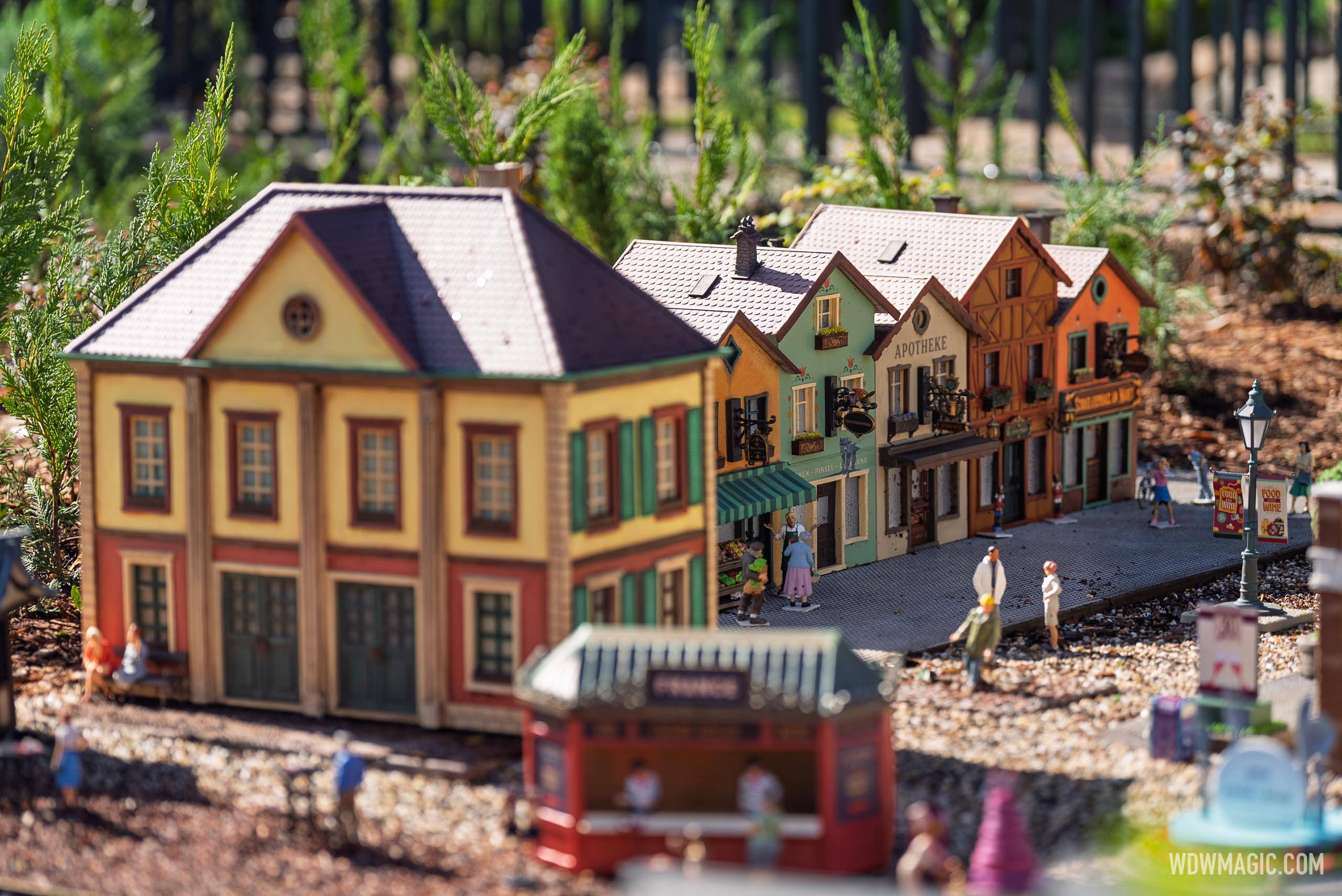 Miniature German village streetscape in the train garden features an Apotheke building, colorful half-timbered facades, a striped awning shop, and tiny figures along a cobblestone street