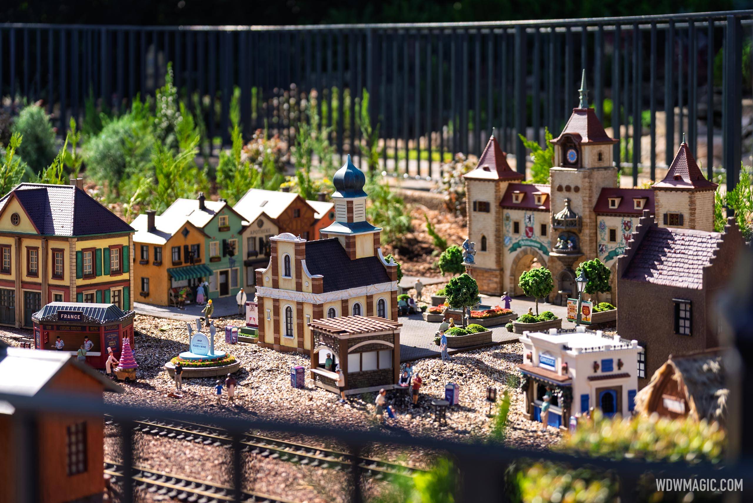 The Germany pavilion train garden overview, showing European-style buildings including a clock tower and church, miniature figures, and a central fountain amid pebbled ground