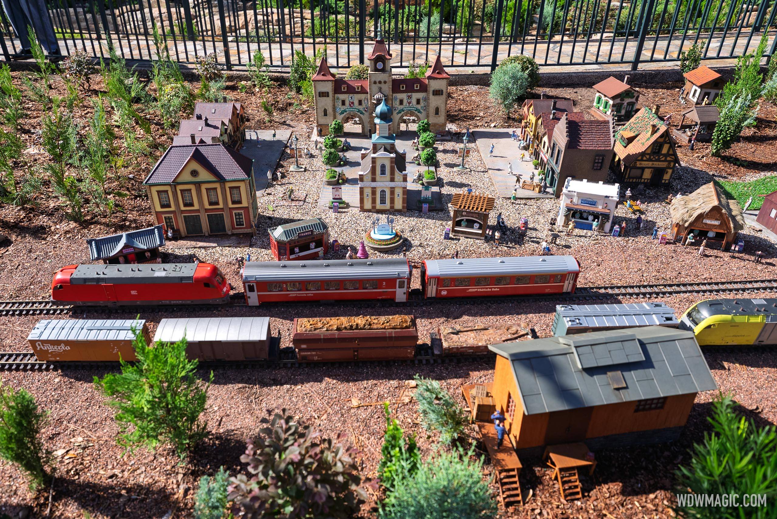 The Germany pavilion train garden features a red Deutsche Bahn locomotive and passenger cars running past a miniature European village with a clock tower, fountain, and tiny figures