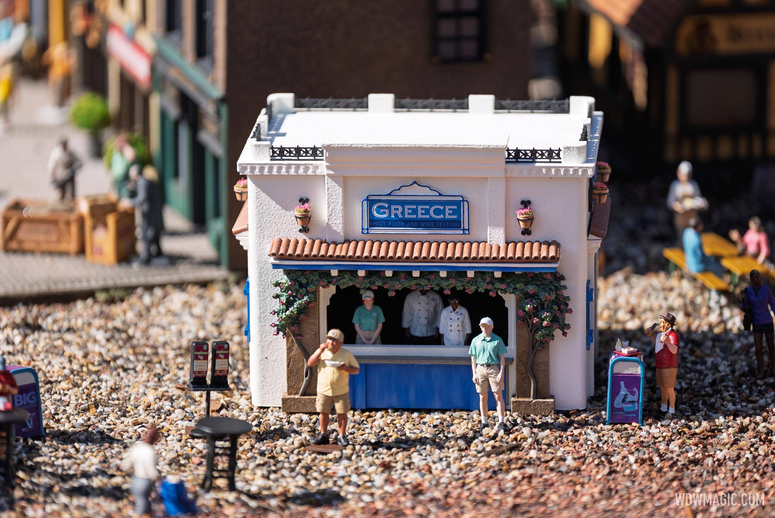 The Greece booth miniature in the Germany pavilion train garden features tiny staff figures behind the counter and guests outside, set against blue-accented Mediterranean architecture