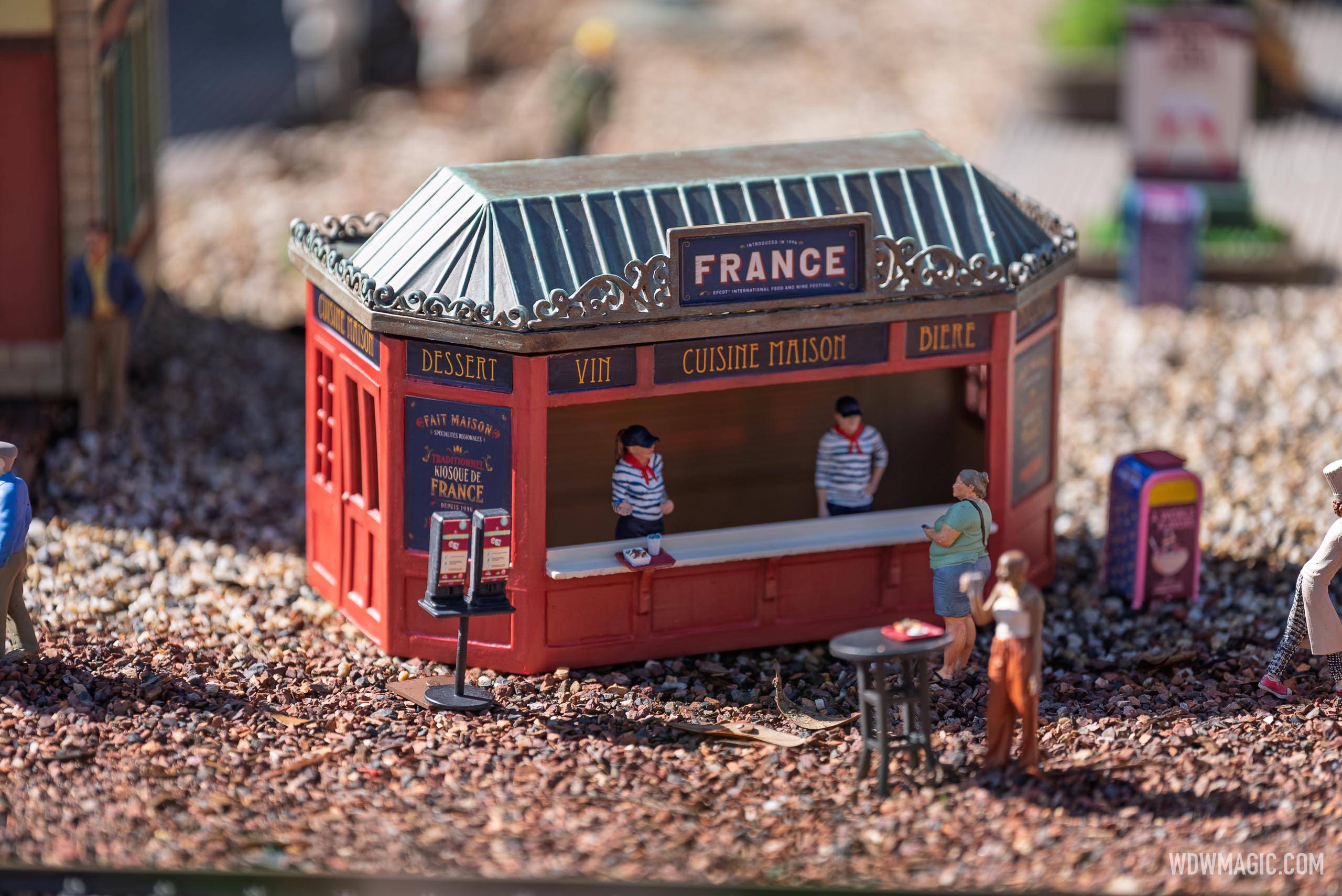 Close-up of the France booth miniature showing staff figures behind the counter and signage for Dessert, Vin, Cuisine Maison, and Bière, with guest figures outside