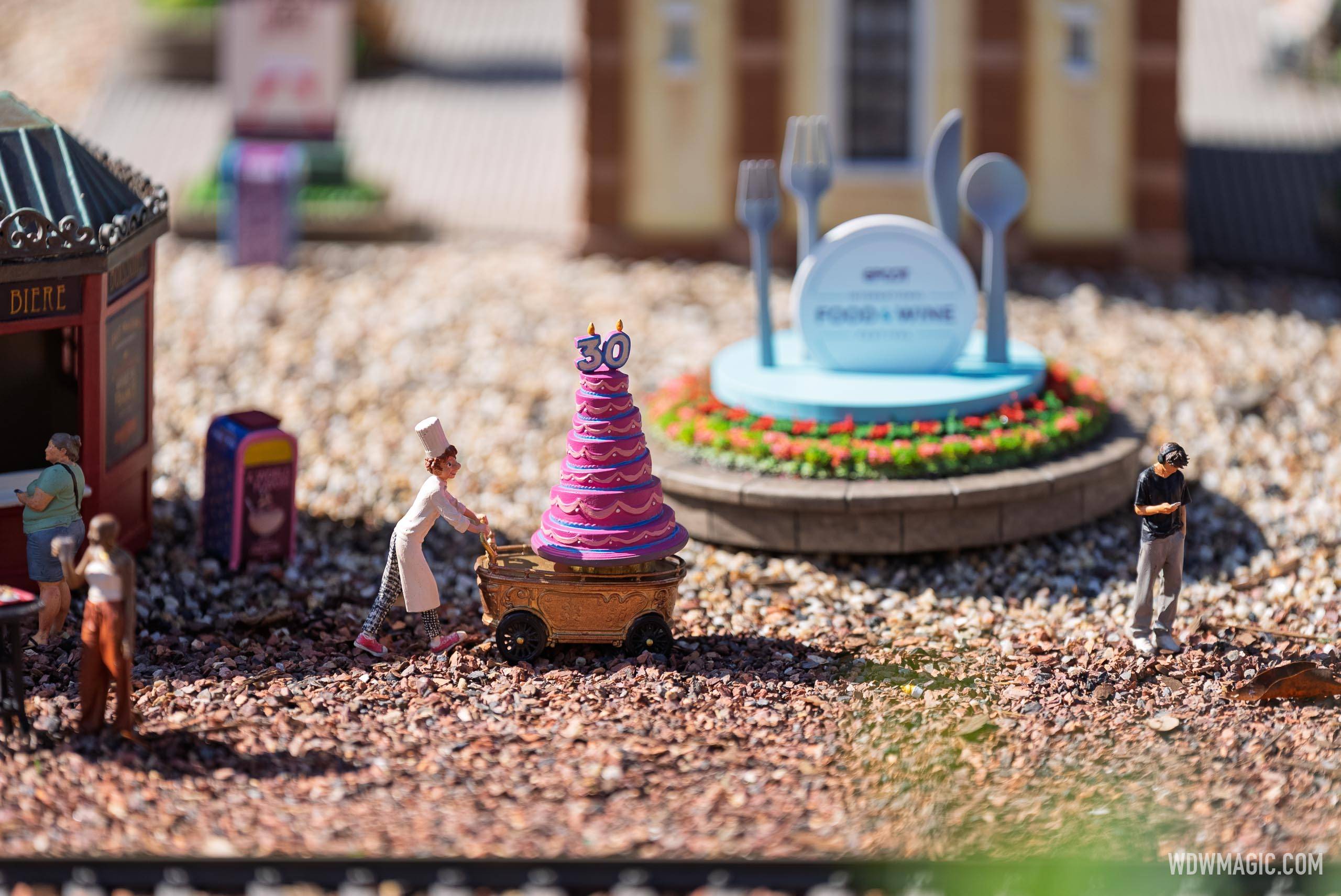 A miniature chef pushes a cart topped with a pink 30th anniversary cake past the EPCOT Food & Wine Festival sign in the Germany pavilion train garden