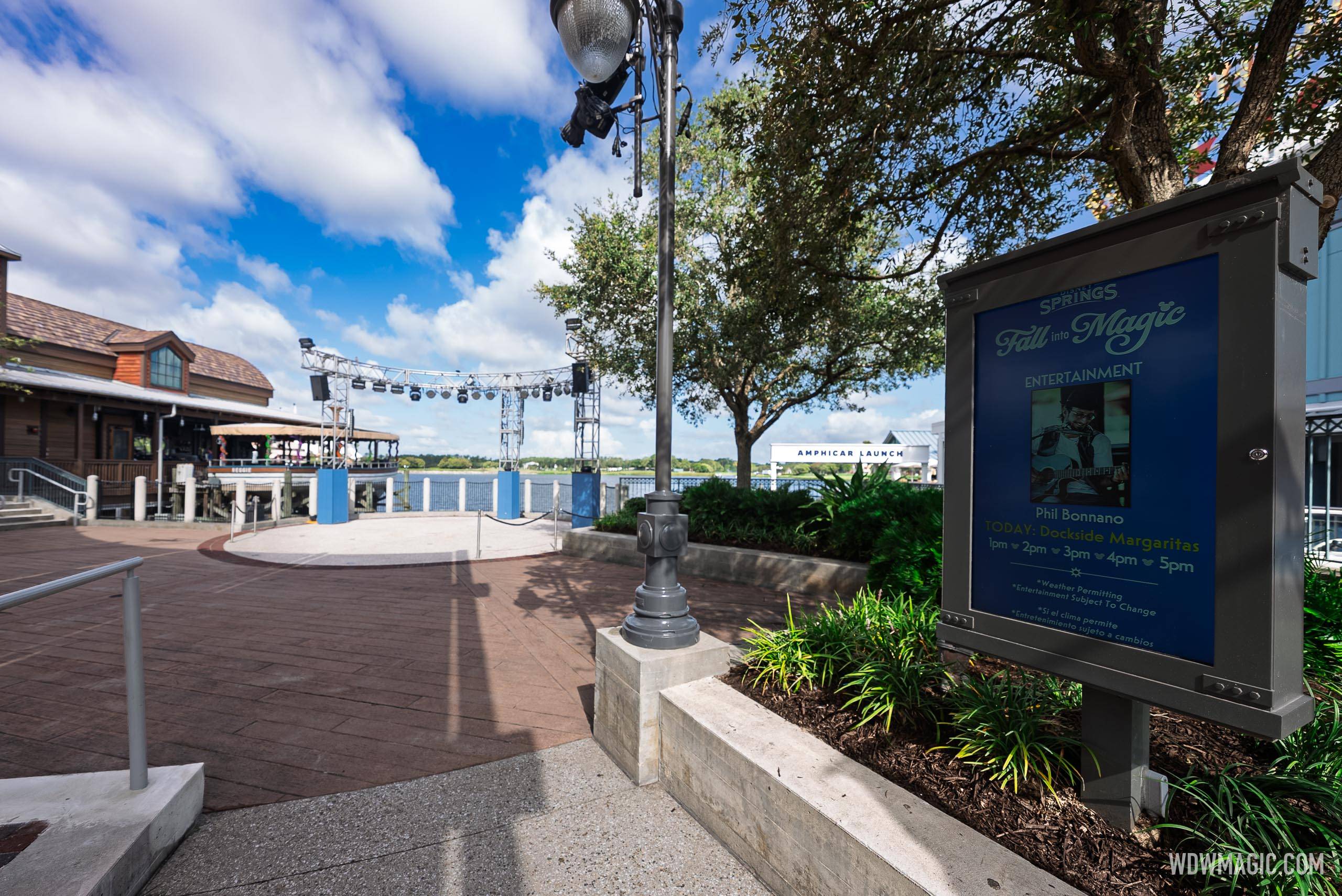 A "Fall into Magic" entertainment sign at Disney Springs advertises Phil Bonnano performing at Dockside Margaritas, with the Americas Landing amphitheater stage visible in the background