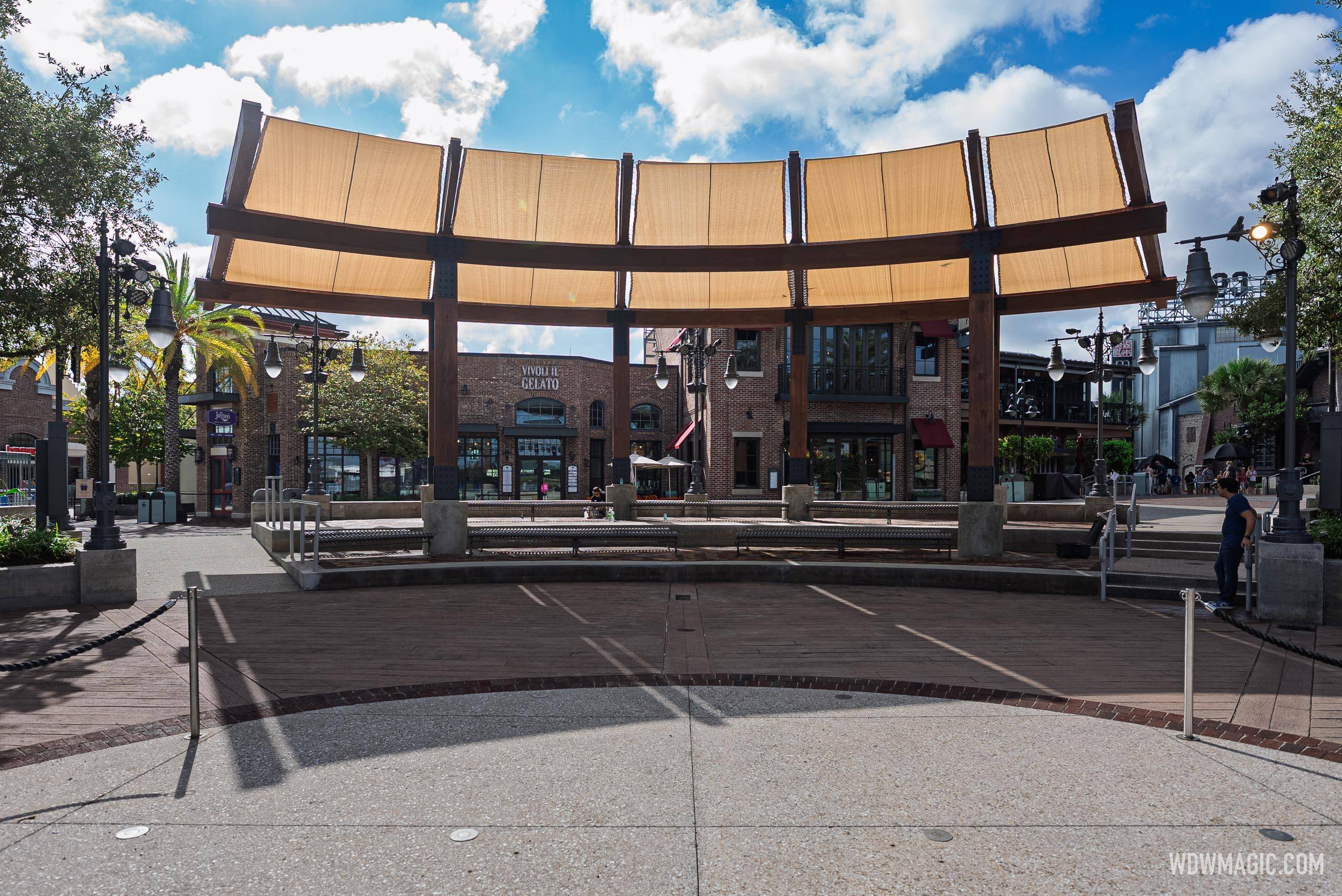 The curved shade canopy at Waterview Park with tan sail panels and dark wood frame, looking toward the Vivoli il Gelato building and surrounding Disney Springs storefronts