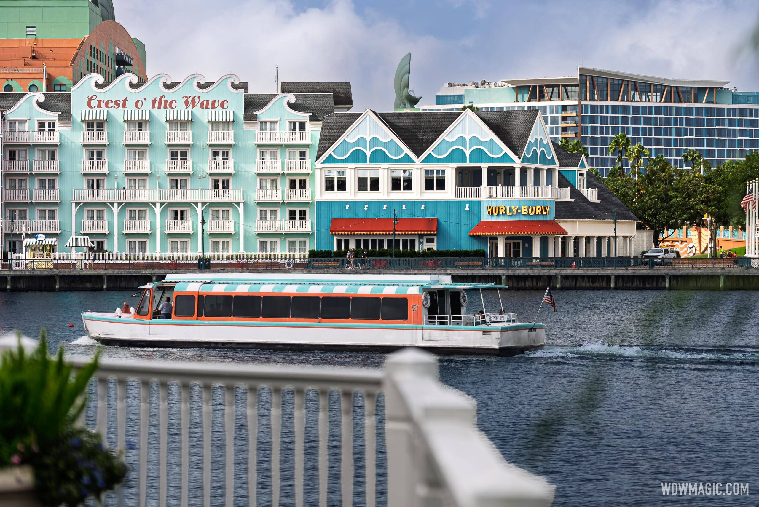 A Friendship boat passes in front of the Hurly-Burly exterior, showing the lit marquee sign, red awnings, and wave-themed teal building alongside the Crest o' the Wave resort facade