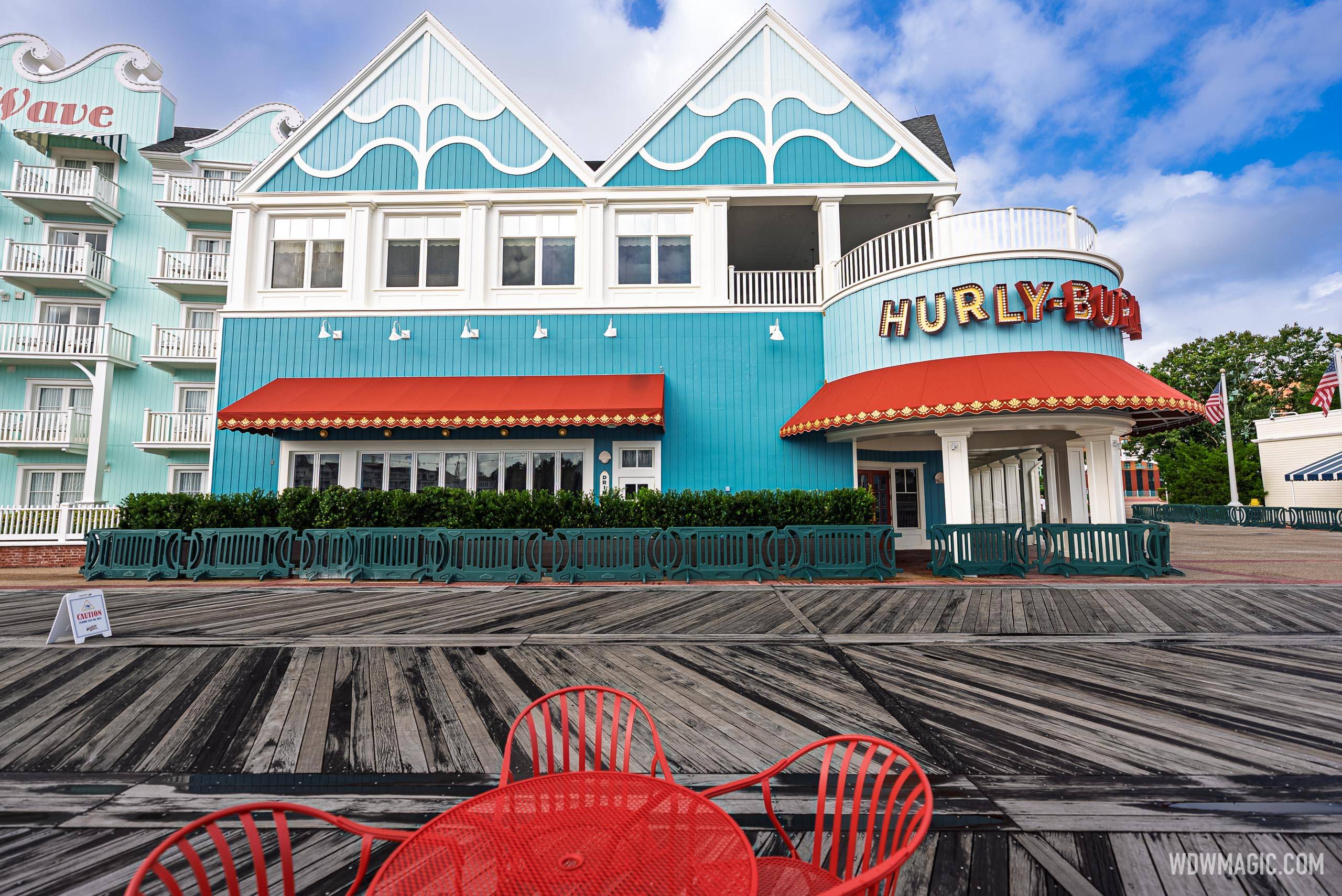 The Hurly-Burly exterior seen from across the BoardWalk, with red awnings, teal wave-themed facade, and lit marquee sign visible alongside red patio furniture in the foreground