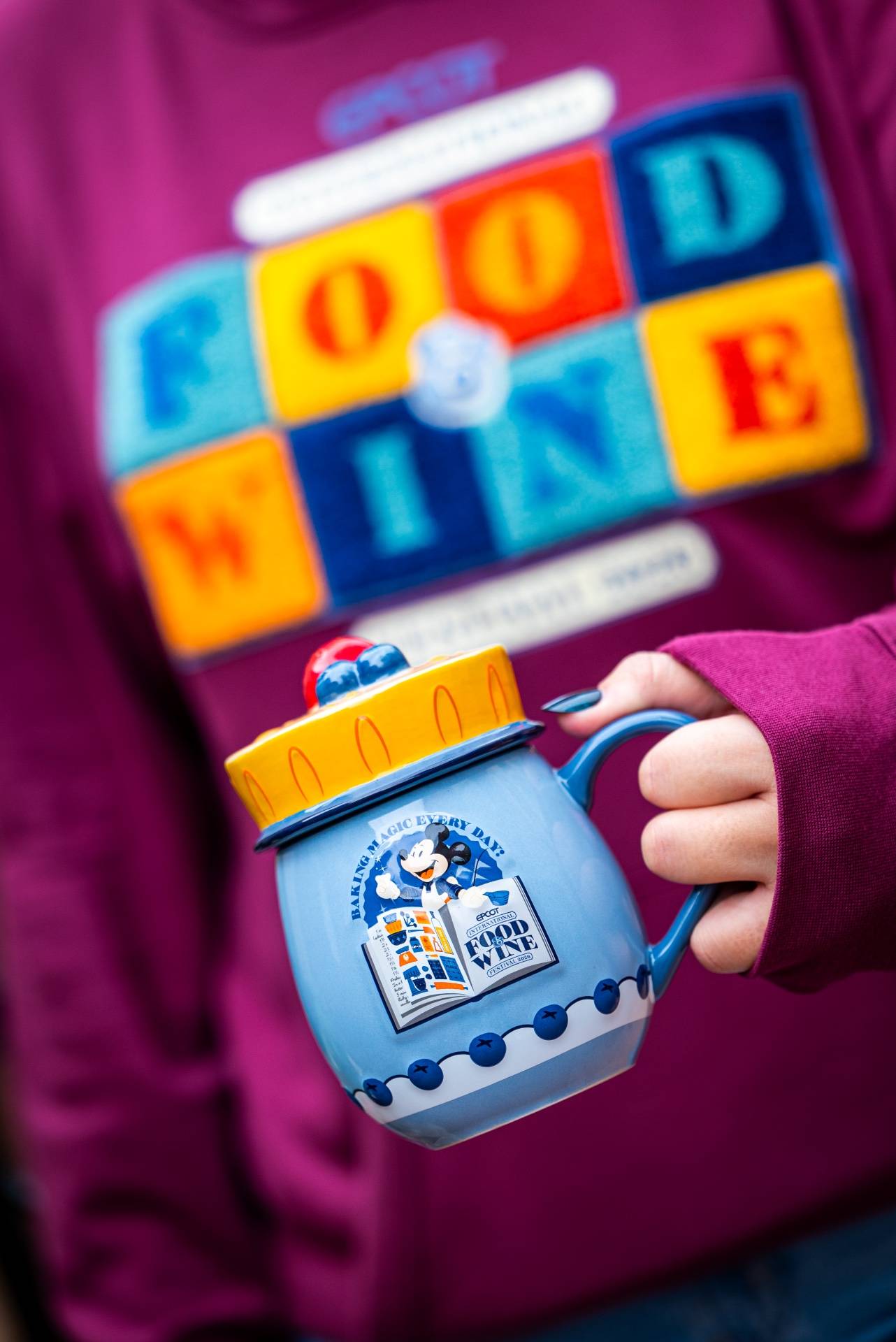 Mickey Mouse "Baking Magic Every Day" mug with yellow lid from the 2026 EPCOT International Food & Wine Festival collection, held against the matching festival hoodie