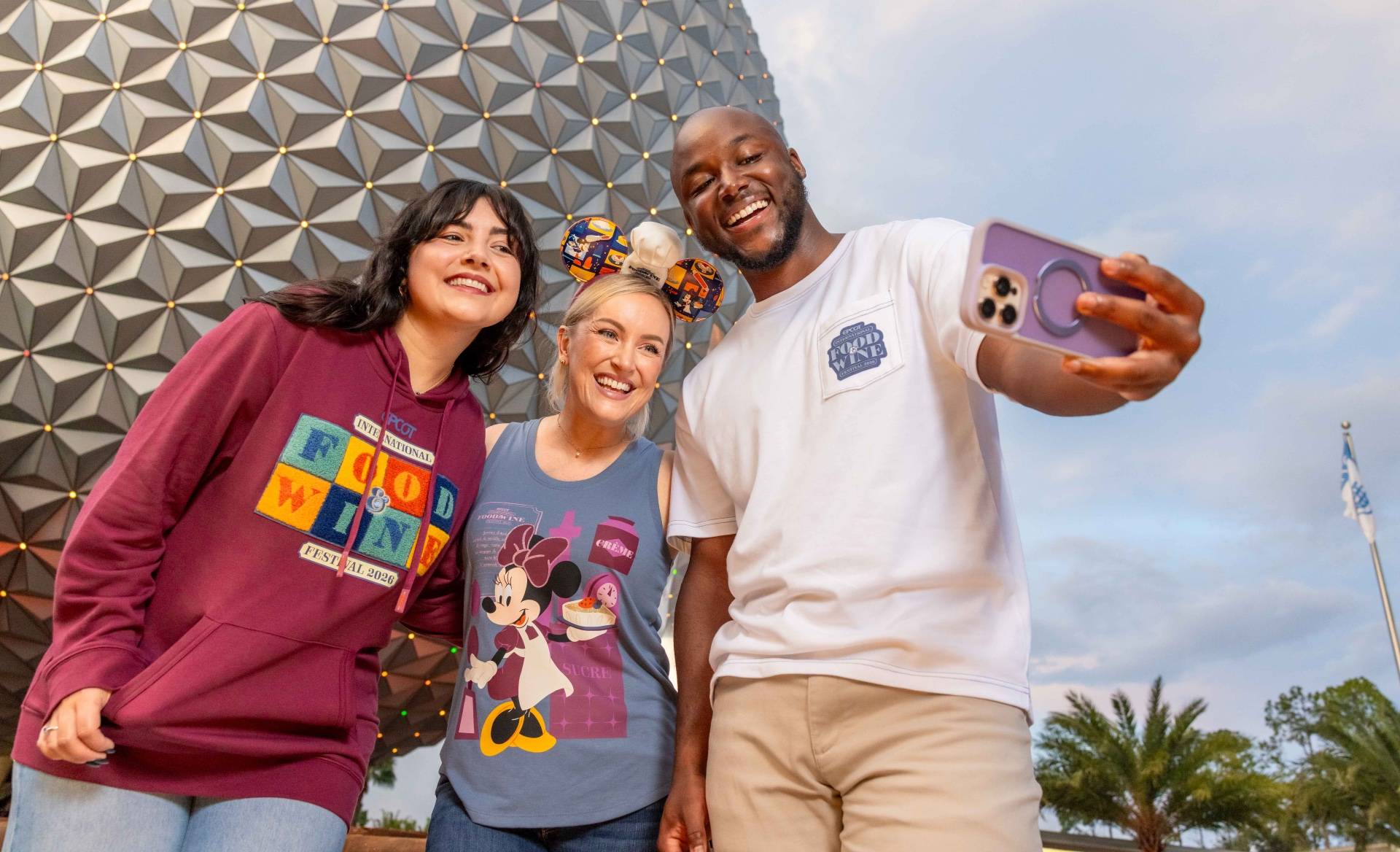 Guests take a selfie in front of Spaceship Earth wearing 2026 EPCOT International Food & Wine Festival merchandise, including the maroon hoodie, Minnie chef ears, and white pocket tee