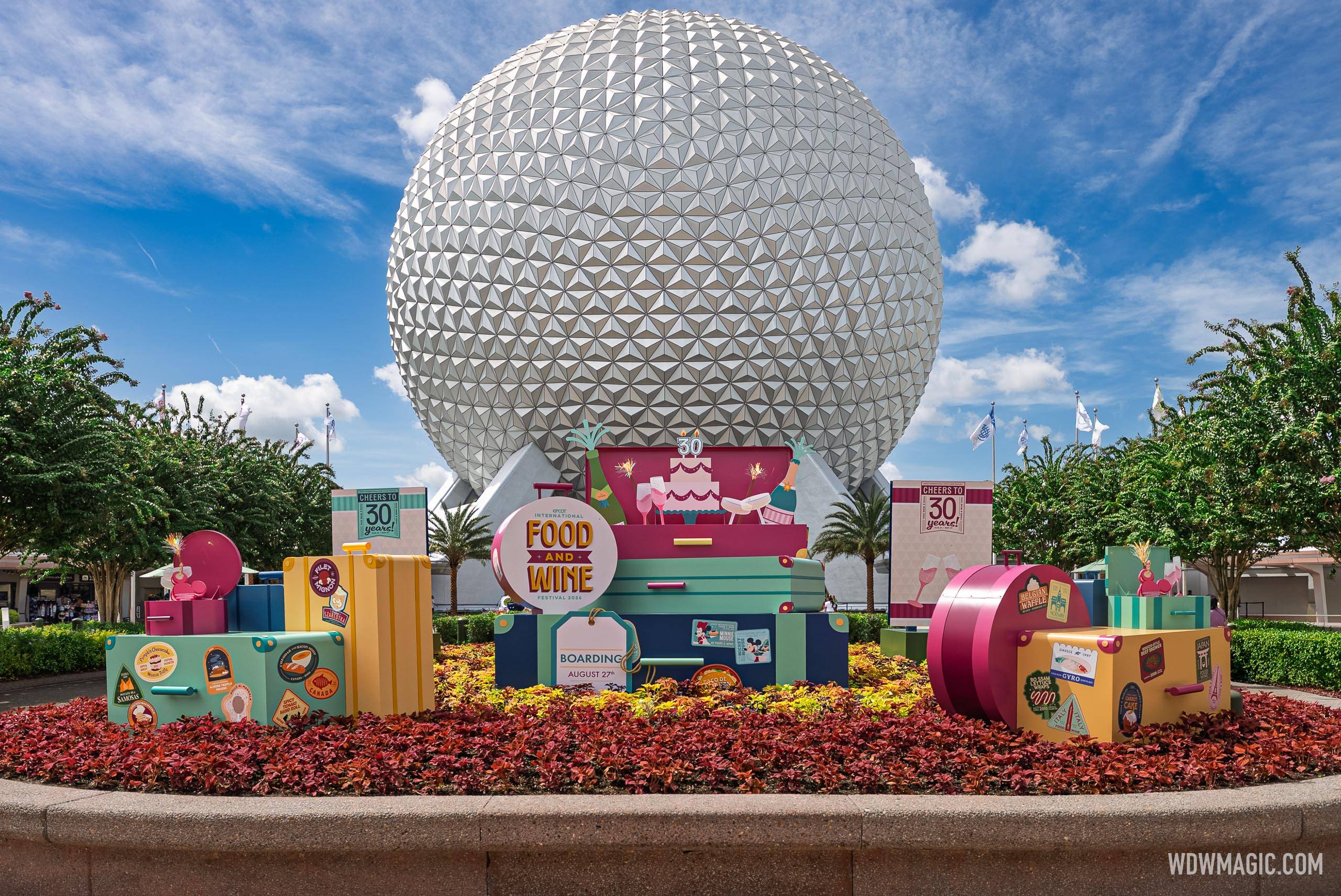 Colorful travel-themed signage and luggage props mark the 30th anniversary of the Epcot International Food and Wine Festival in the Spaceship Earth flower bed