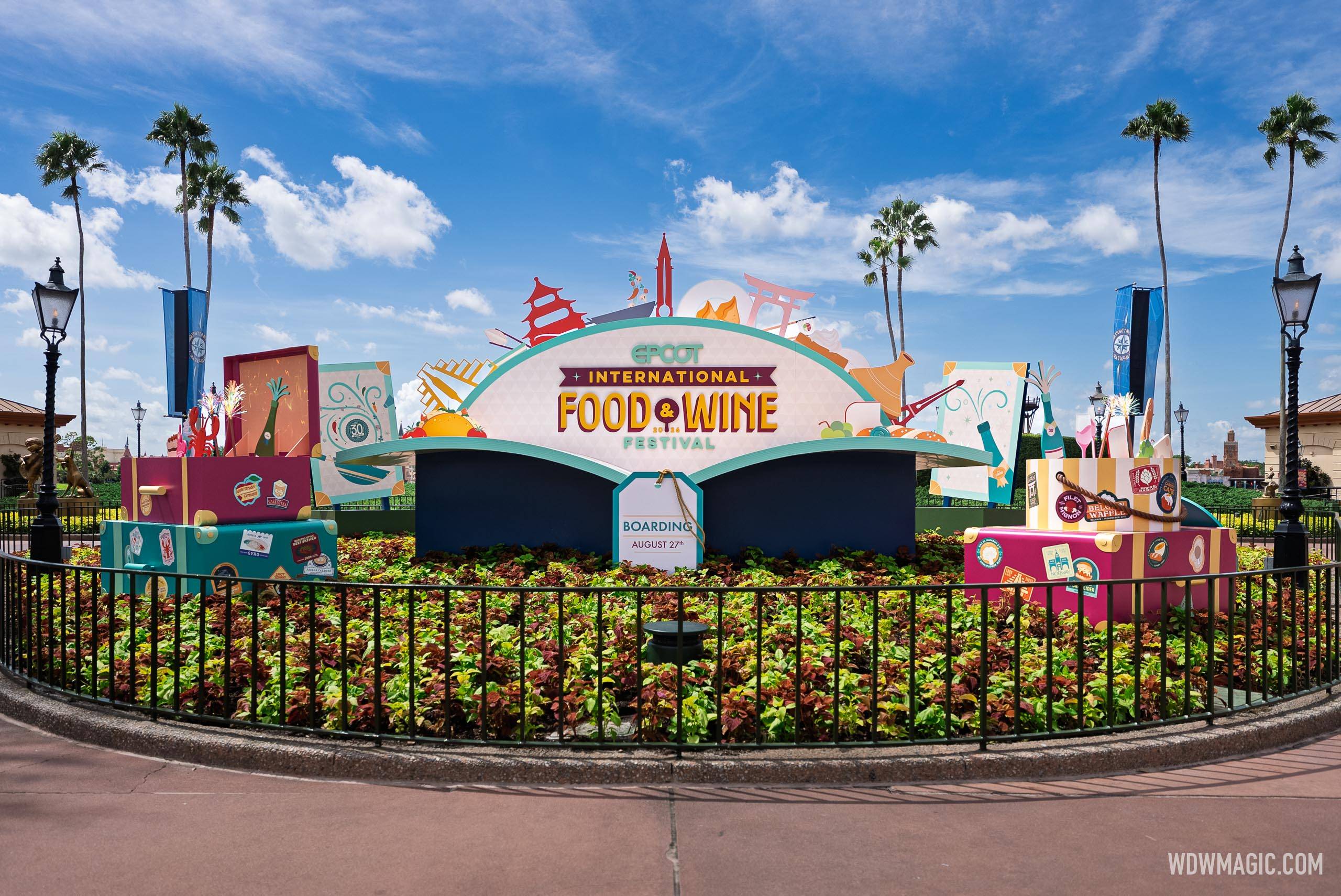 The Epcot International Food and Wine Festival World Showcase Plaza sign with travel-themed luggage props sits in the World Showcase Plaza flower bed, with a "Boarding August 27" tag displayed on the sign