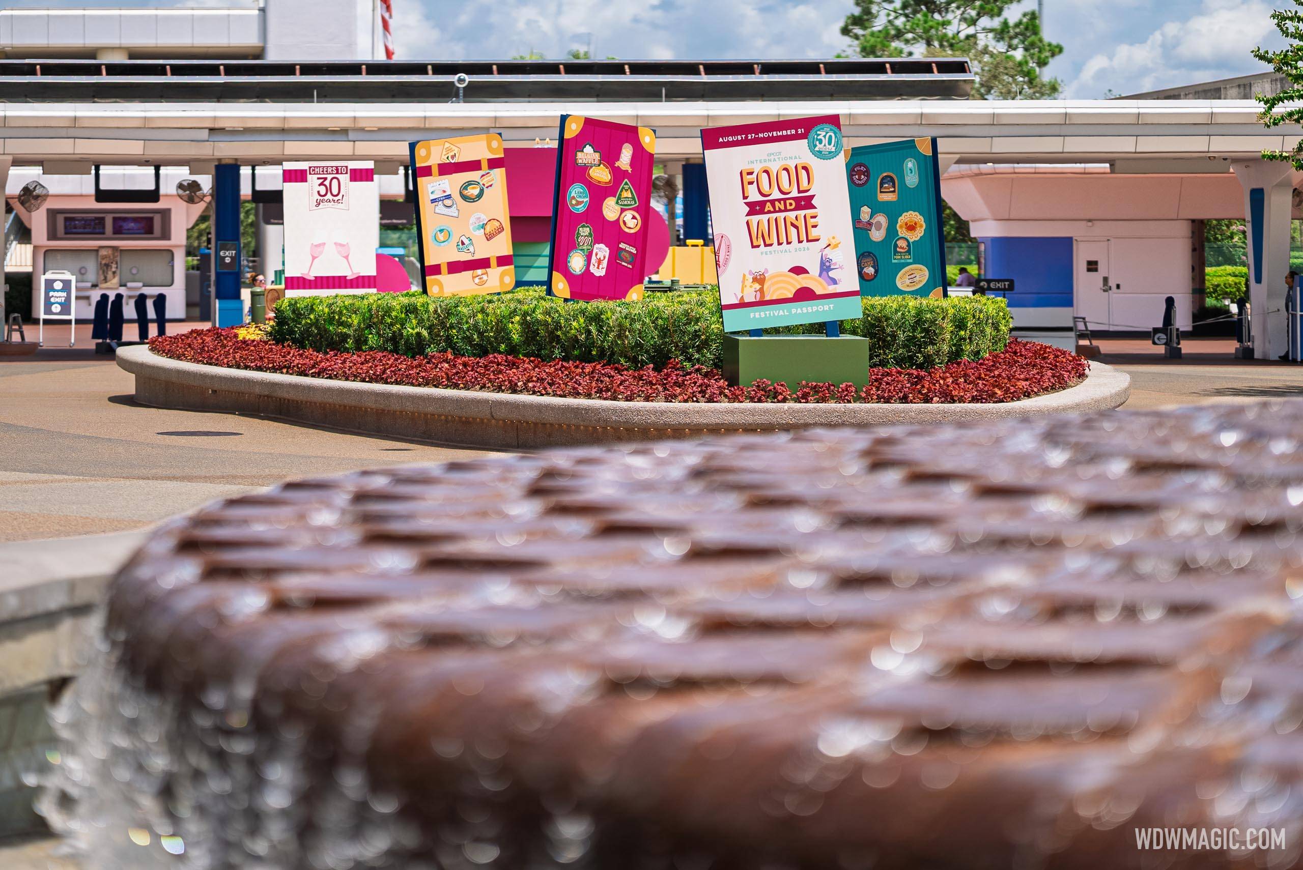 Festival Passport and "Cheers to 30 Years" signage displayed in the Spaceship Earth flower bed, photographed from the fountain at the Epcot main entrance plaza