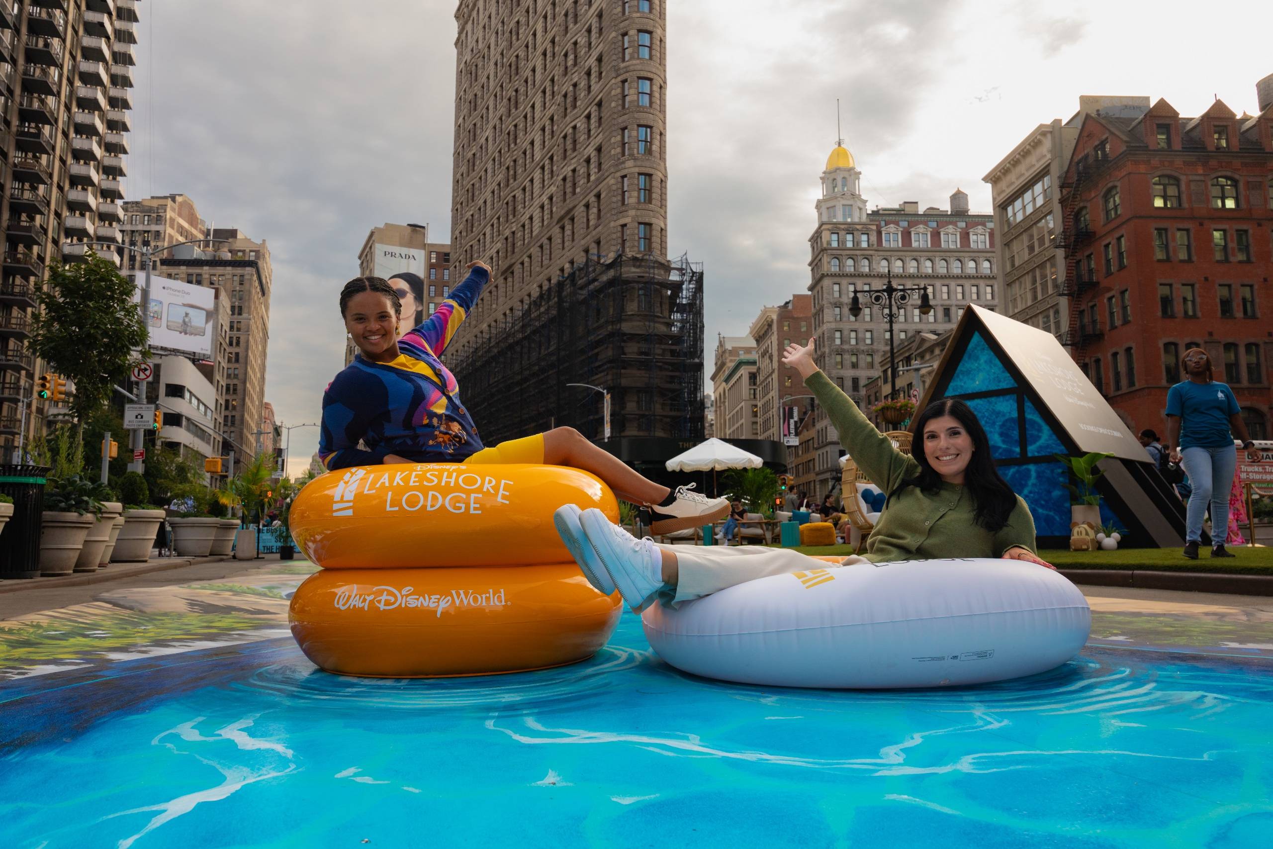 Two guests pose in Lakeshore Lodge-branded inflatable rings on the 3D Daydream River mural at Flatiron Plaza, with the Flatiron Building visible in the background