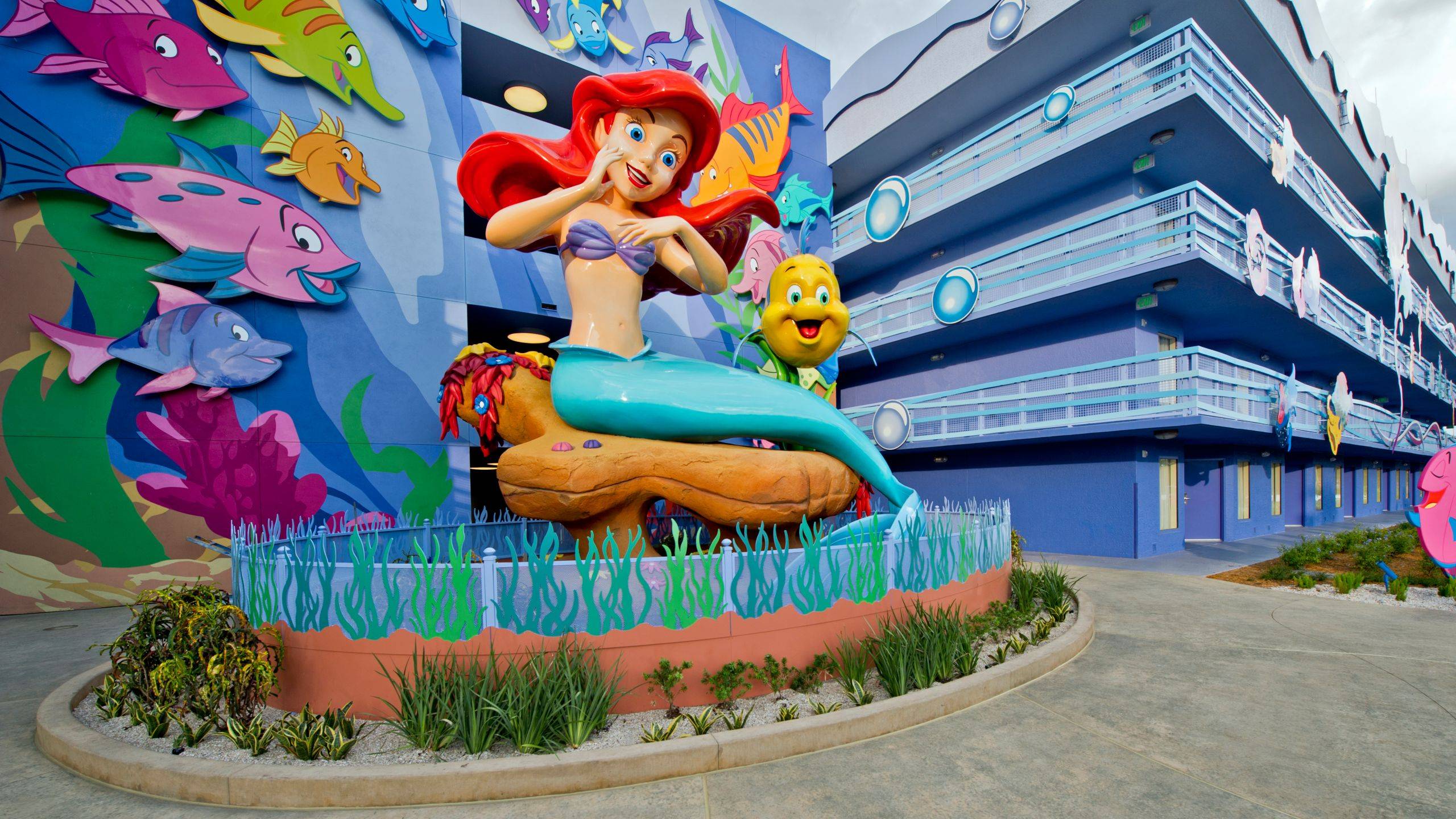 Large Ariel and Flounder statues anchor the Little Mermaid section courtyard, surrounded by colorful fish sculptures and undersea-themed building murals
