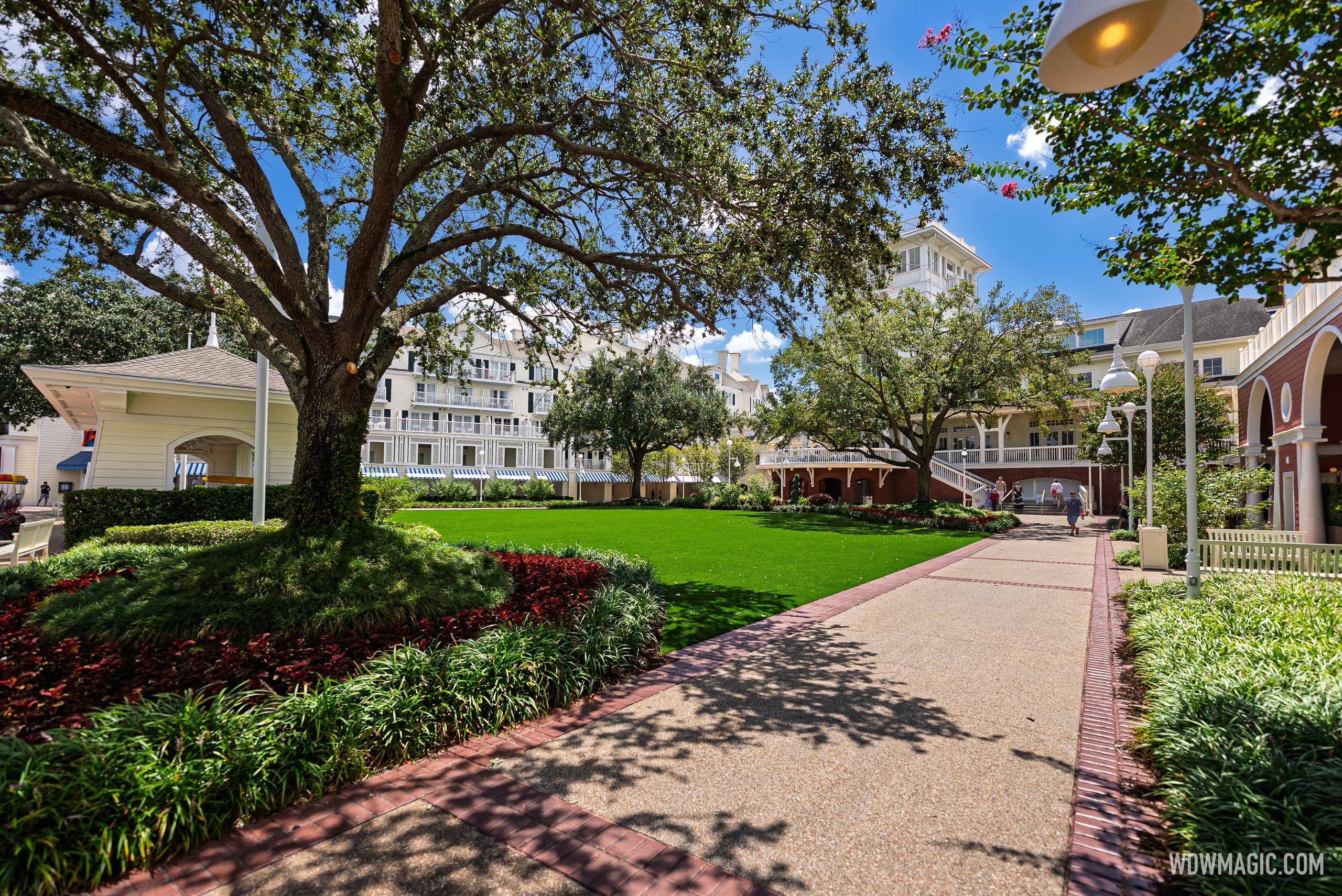 The Village Green courtyard pathway at Disney's BoardWalk Inn, flanked by manicured gardens and newly installed artificial turf with the resort's main building visible in the background